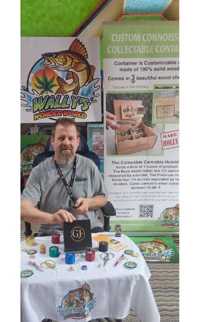 Greg from Wally's Wonder World sitting at a trade show display table presenting custom wooden cannabis humidors, grinders, and accessories.